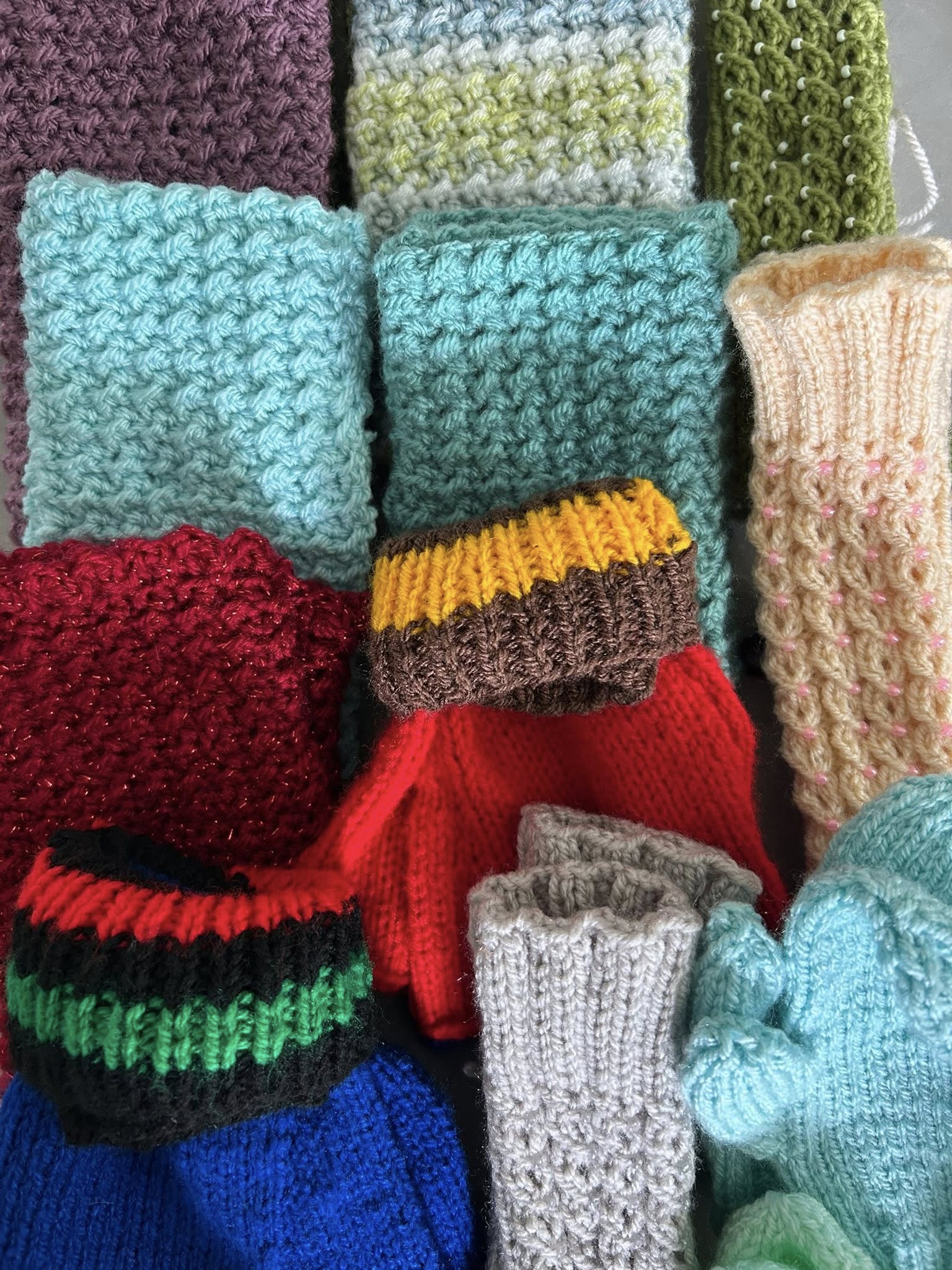 Close‑up of assorted hand‑knitted accessories in mixed colours and textures, including mitts, socks and patterned swatches.