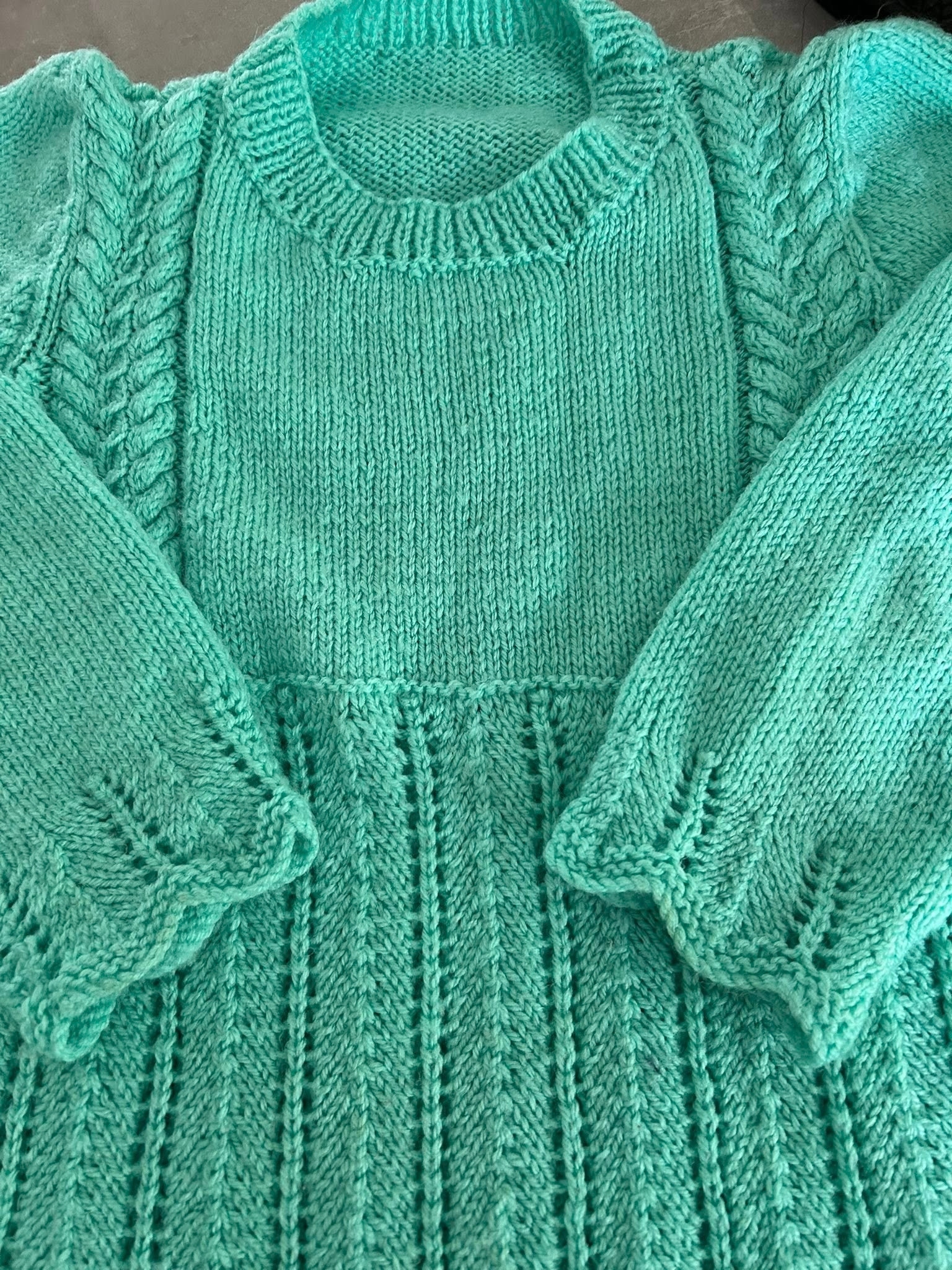 Close‑up of a child’s hand‑knitted turquoise jumper showing textured stitches, ribbing and scalloped sleeve edges.